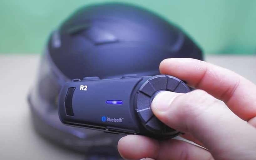 Airide R2 Motorcycle Bluetooth Headset with blue indicator light, ready for Bluetooth connectivity, and a black Sedici Strada II helmet in the background.