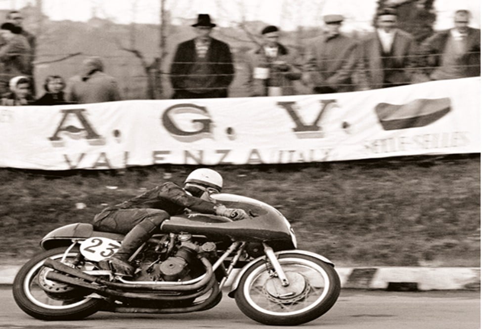 A racer leans into a tight corner, as an AGV advertisement banner looms in the background. Several spectators can be seen standing behind the banner.