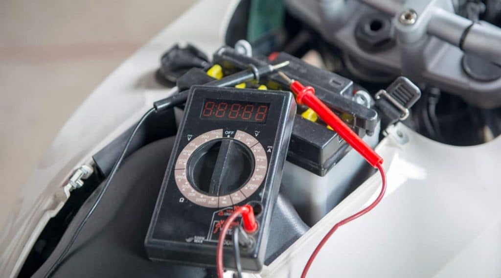 A multimeter rests on a motorcycle battery which is still in its enclosure but disconnected from the bike’s accessories.