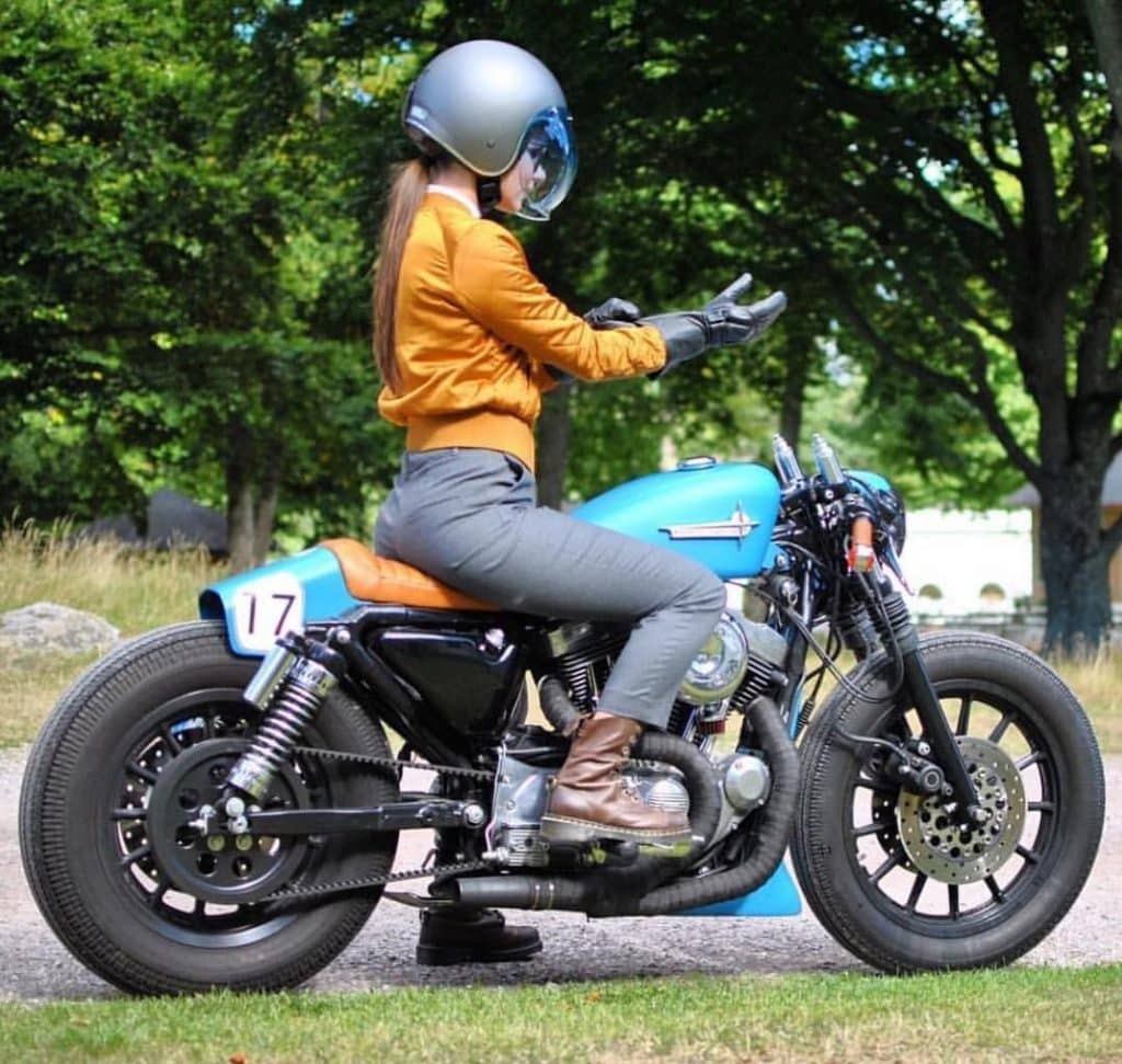 A lady rider in thick brown boots, casual gray pants and orange jackets sits on a blue giant V-twin powered cafe racer. The motorcycle has heavy set tires, belt final drive and shortened exhaust pipes wrapped in some blackened material. The front fender has been removed and the rear one is shortened and integrated into the seat. Huge shocks in the rear support a slim swingarm and we have single and dual-caliper disc brakes in rear and front respectively. Handlebars are clip-ons and drooping downward and outward.