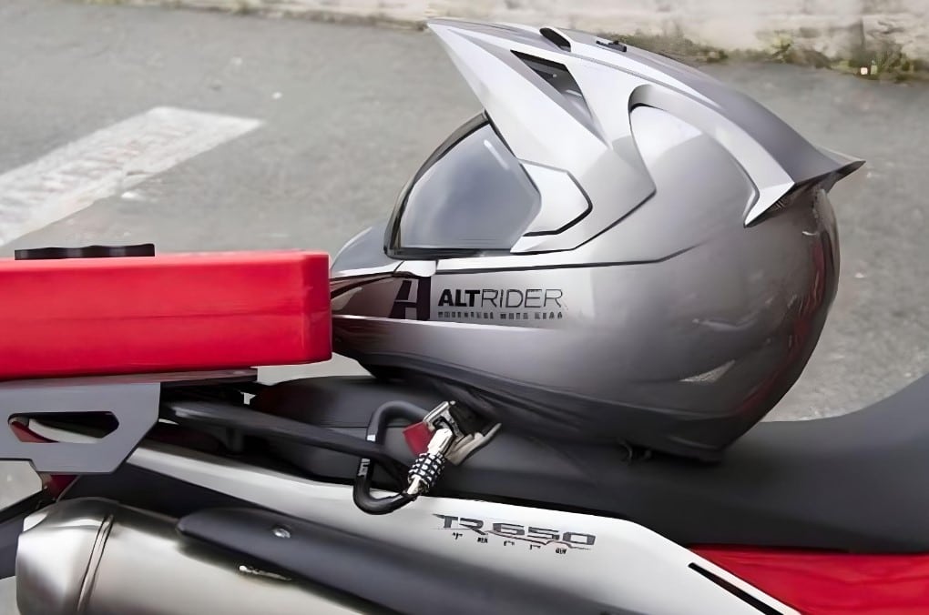 A four combination  HelmetLok 4104 Carabiner Style Lock secures a silver dual sport helmet with AltRider decor to the grab-bar of a Husqvarna TR650 Terra.