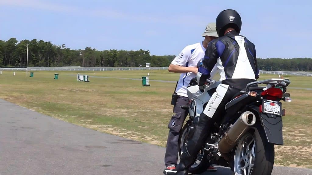 A white t-shirt-clad instructor guides a trainee wearing a 16-year AGVSPORT’s leather suit and a helmet adorned with a DOT sticker at the back at Keith Code's California Superbike School. While motorcycle leathers can last for 15-20 years or even longer with proper care, you should replace a helmet after every 5 years.