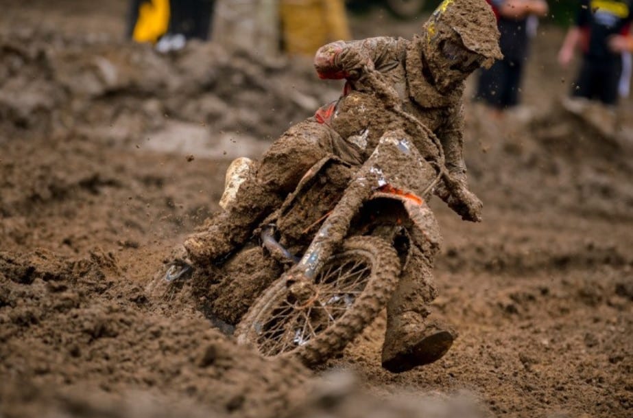 A motocross rider dressed in full gear kicks up mud while racing on a track, with spectators in the background.