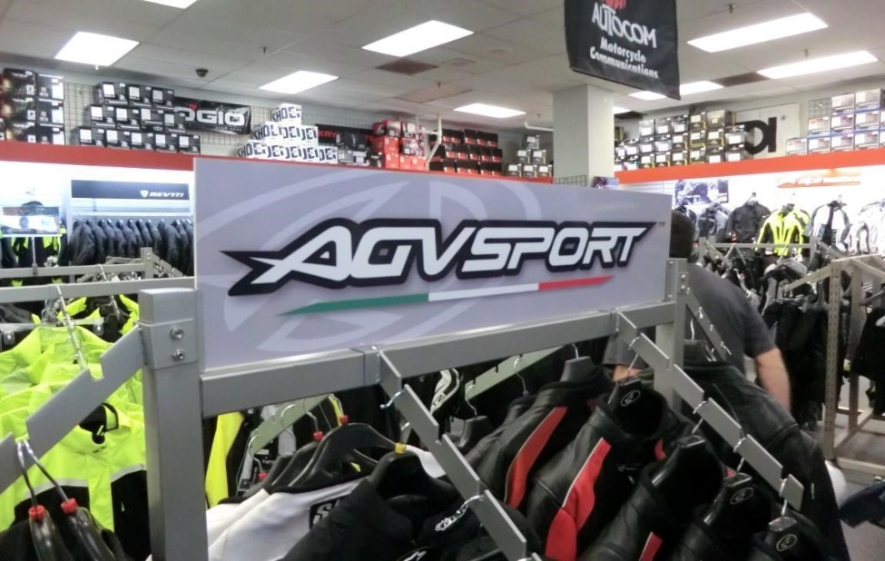 The AGVSPORT apparel is neatly displayed on a clothing rack in-store, with the AGVSPORT name design board placed prominently on top of the rack.