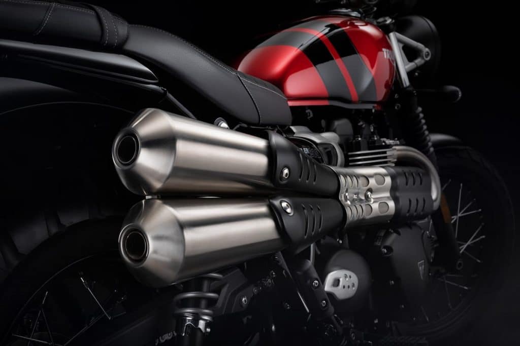 A closeup of the up-swept dual exhaust new Triumph Scrambler 900. It features a distinct bench seat, chunky gas tank, tall suspension, and spoked wheels.