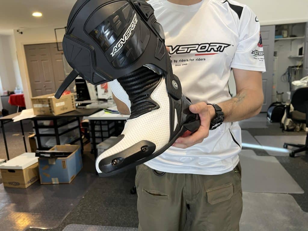 Denis displays the AGVSPORT Monza race boot, showcasing ventilation perforations in the white section, protective toe sliders on the front-right, and armored heel plates at the back for enhanced safety and breathability.