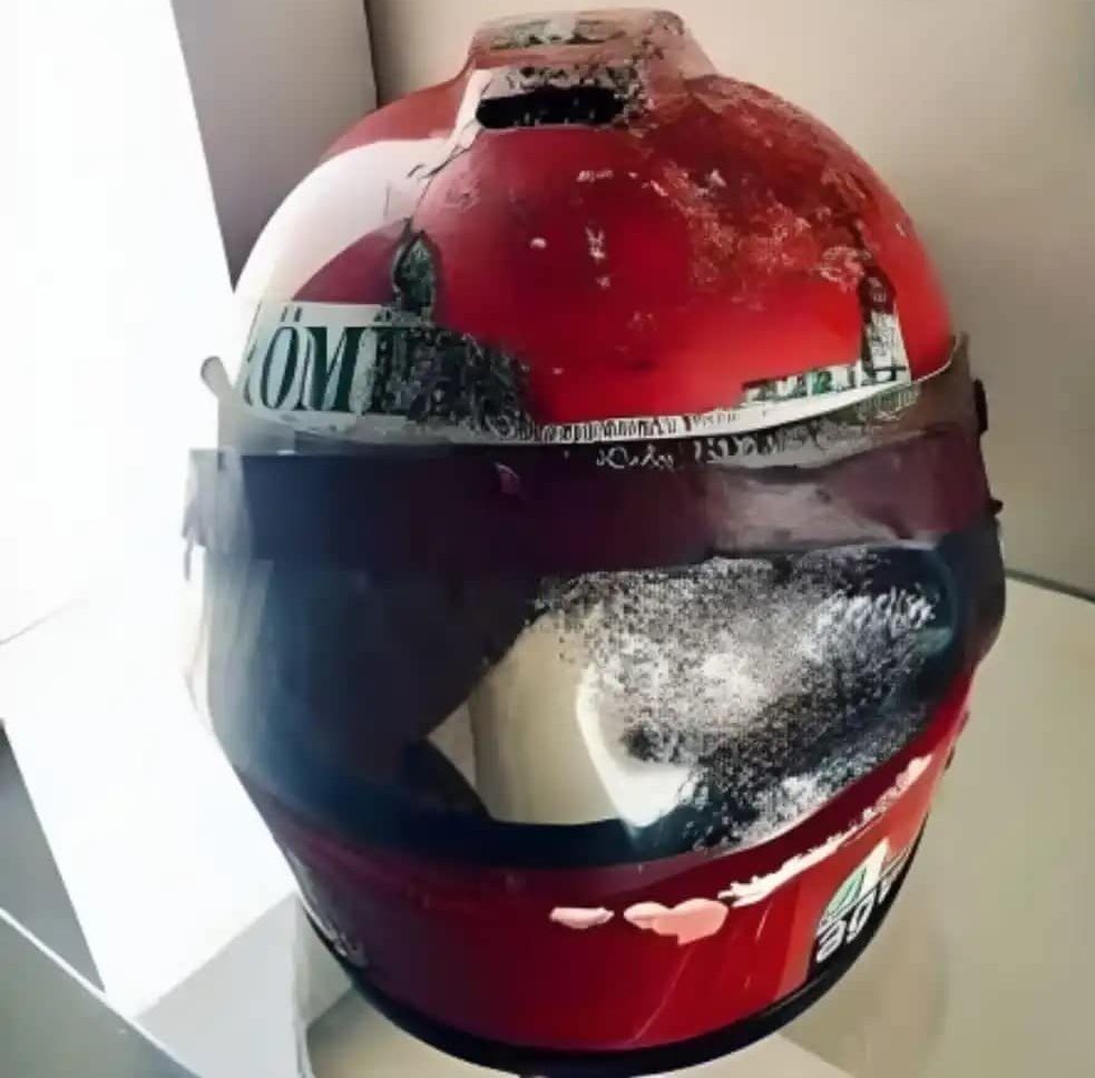 Displayed on a glass case in my AGV Sports Group office in Maryland, United States, the AGV X1 helmet highlighting a vibrant red design adorned with 'Niki Lauda' in crisp white lettering on both sides, alongside logos for AGV, Goodyear, and Romerquelle (partial logo obscured). A faint mark is evident on the front of the chin guard where a Raiffeisen Bank sticker once resided.
