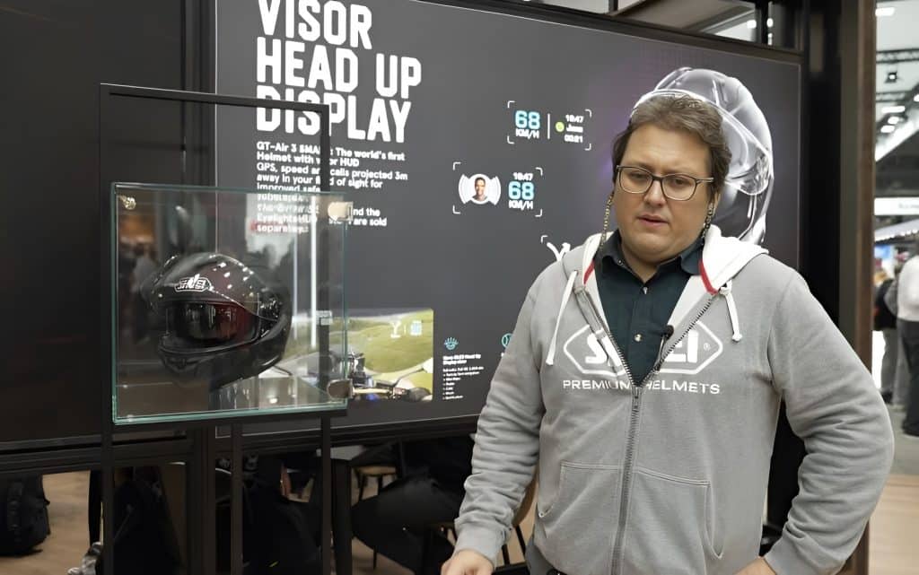 Fabian Naziruka stands next to a glass display case containing the the Shoei GT-Air 3 Smart helmet with EyeLights AR display. Behind him, a large screen reads "Visor Head Up Display" as he explains to me how the EyeLights HUD system will enhance rider safety and situational awareness by reducing glance-away time, delivering an intuitive low-distraction interface, and creating a scalable, connected platform that can add navigation, telematics and advanced alerts in future updates.
